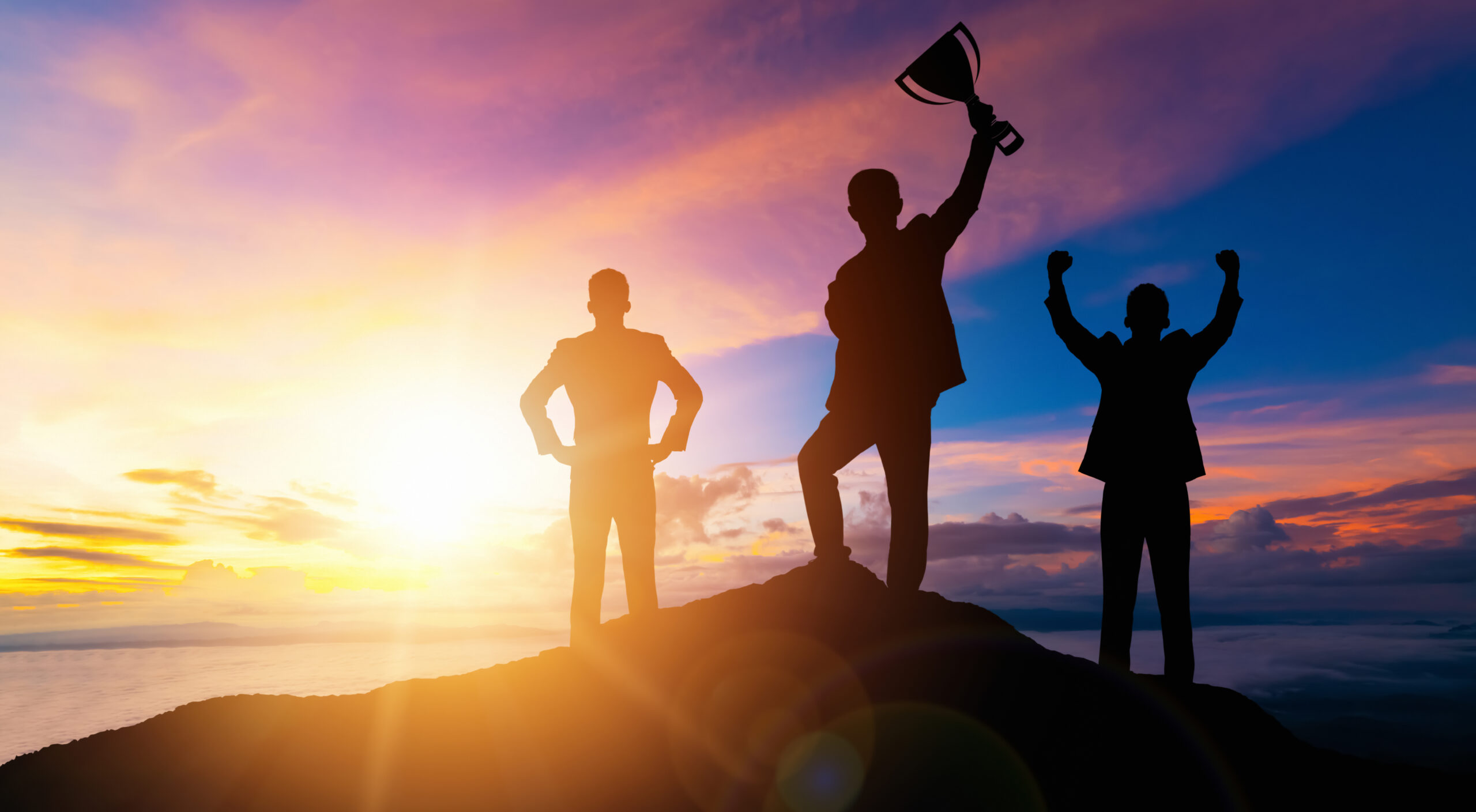 Three silhouette figures on a mountain top in front of a sun set holding a trophy to signify victory